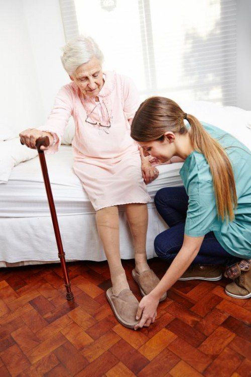 Altenpfleger hilft Seniorin beim Aufstehen und Anziehen der Hausschuhe im Pflegeheim