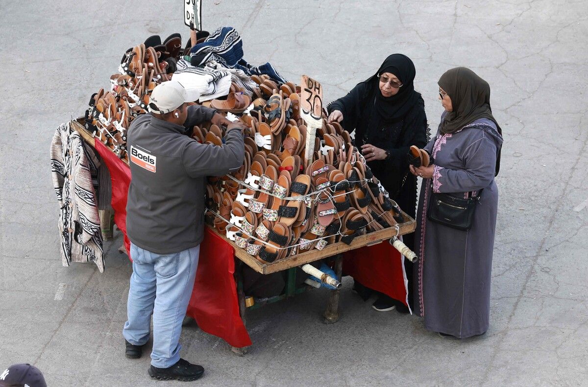 Ein Schuhverkäufer in Marrakesch mit Kundinnen auf dem UNESCO-gelisteten Platz Jemaa el-Fna im Herzen der Medina.